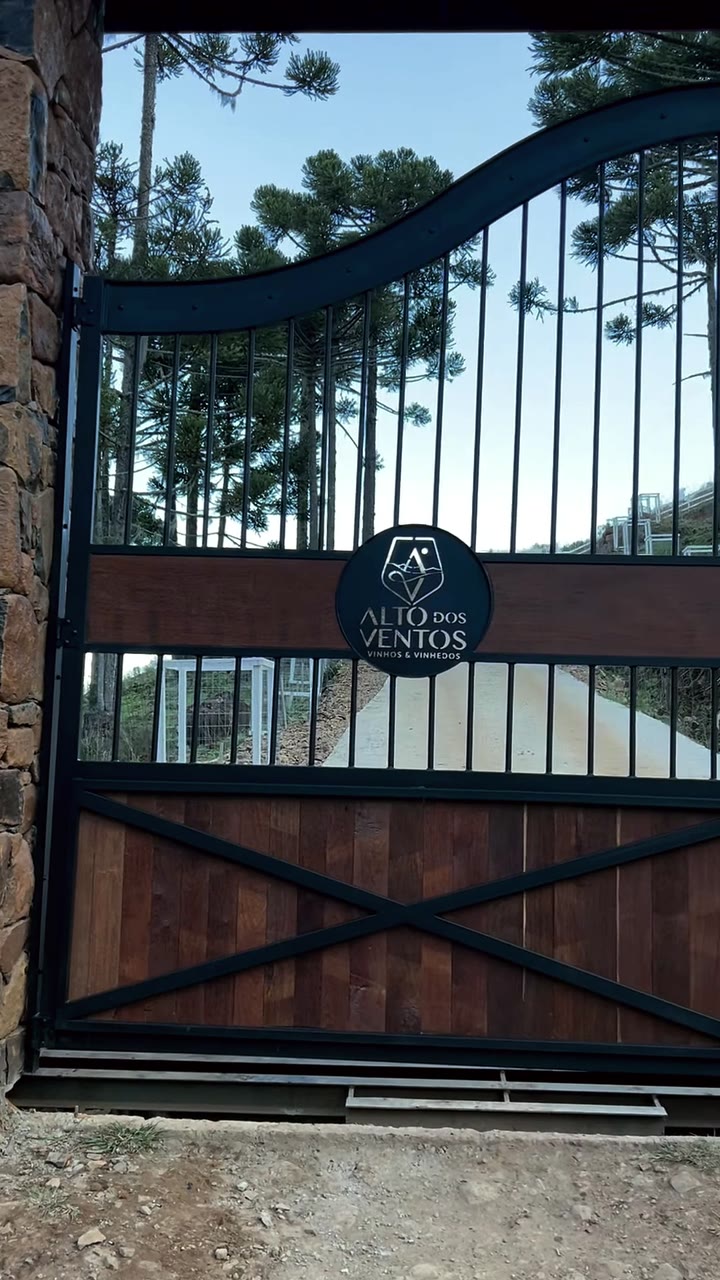 Portão Serrano em aço escuro e madeira, com brasão personalizado ao centro, entre pilares de pedra e araucárias.